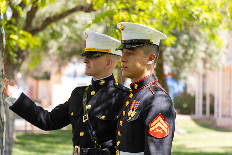 Two individuals dressed in Naval ROTC commissioning uniforms standing outside among trees.